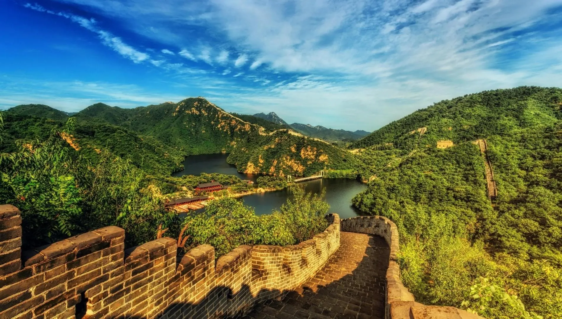 View from the Great Wall of China, showcasing surrounding mountains, lush trees, and serene lakes in the landscape below.