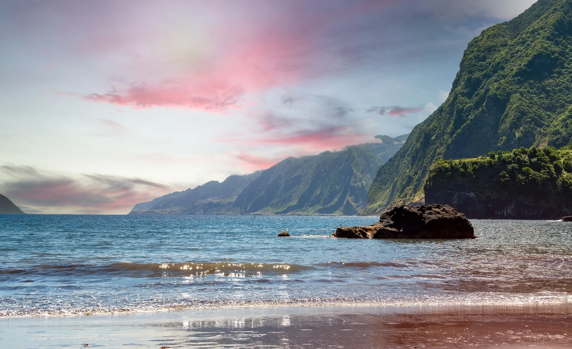 View from the beach at sunset, showcasing the coastline of Madeira with the sun casting a warm glow over the ocean and cliffs.