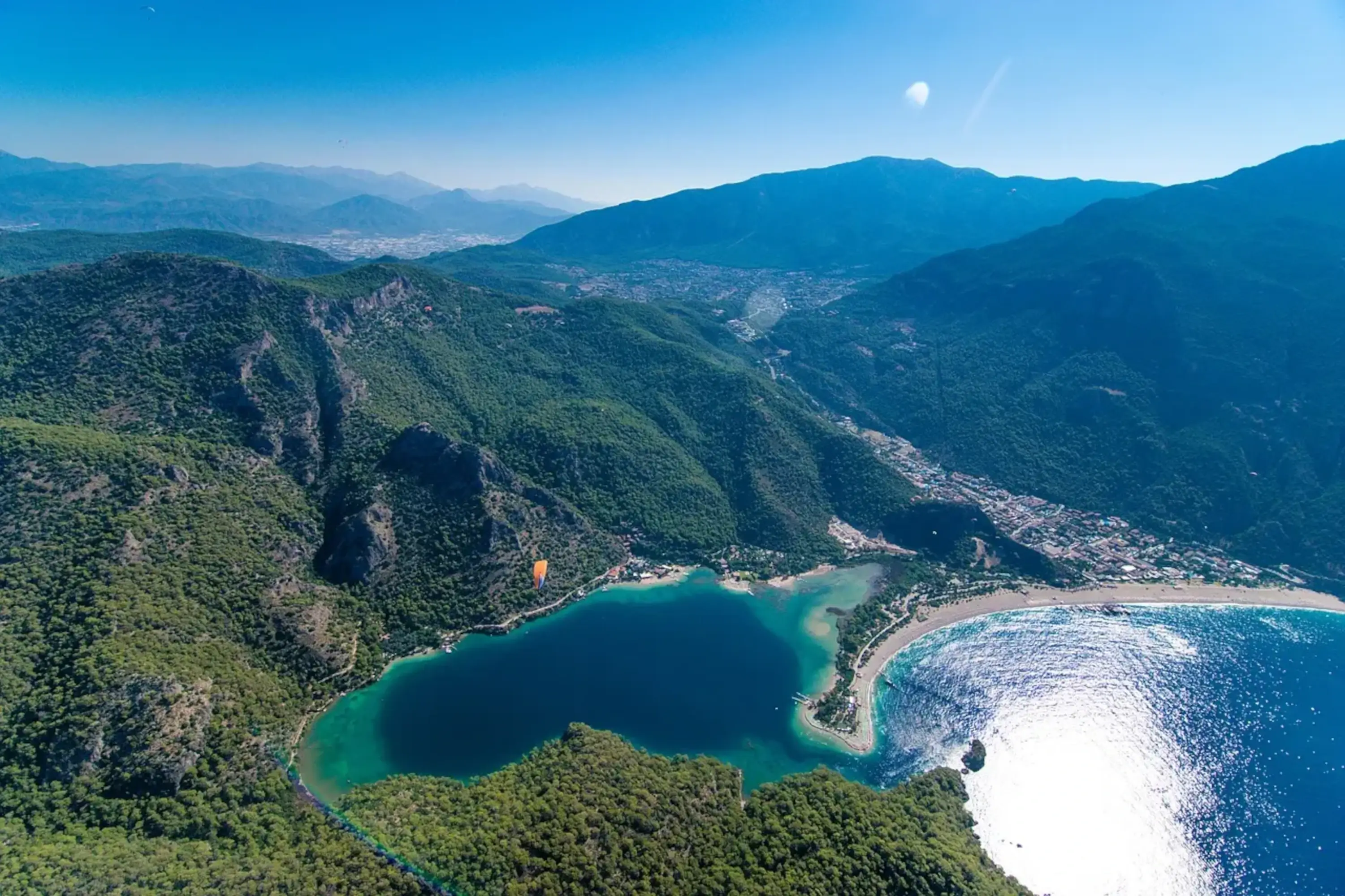 Aerial view of the Ölüdeniz coast in Turkey, featuring a sandy beach, turquoise waters, and the surrounding mountainous landscape.